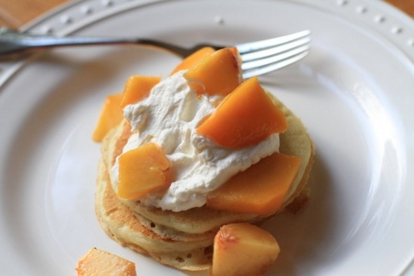 A pancake stack on a white plate, topped with peaches and whipped cream.