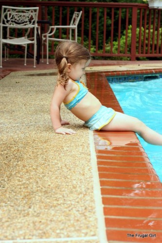 A little girl in a blue bikini, sitting on the edge of a pool.