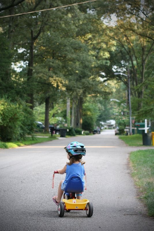 A little girl with a helmet on, riding a tricycle down a tree-lined stree.