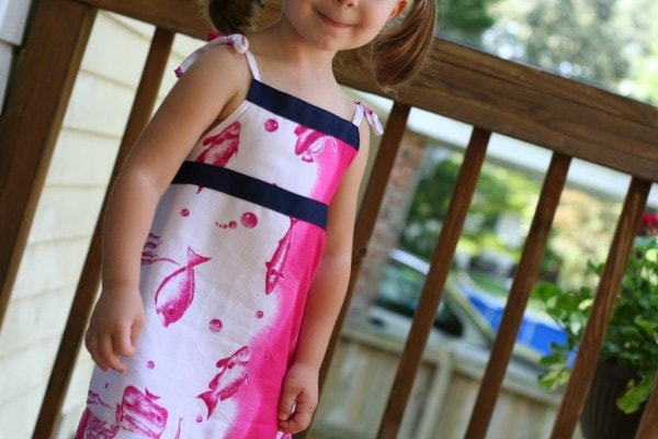 A little girl wearing a white and pink fish dress with navy blue ribbon trim.