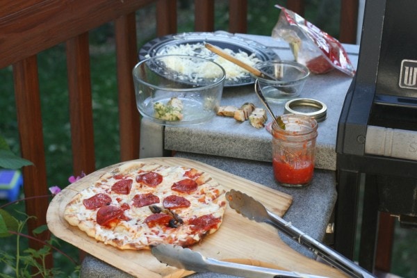 A view of a grill with pizza-making ingredients on the side shelf.