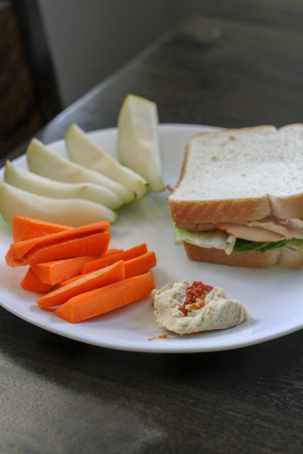 Turkey sandwich on a plate with produce and hummus.