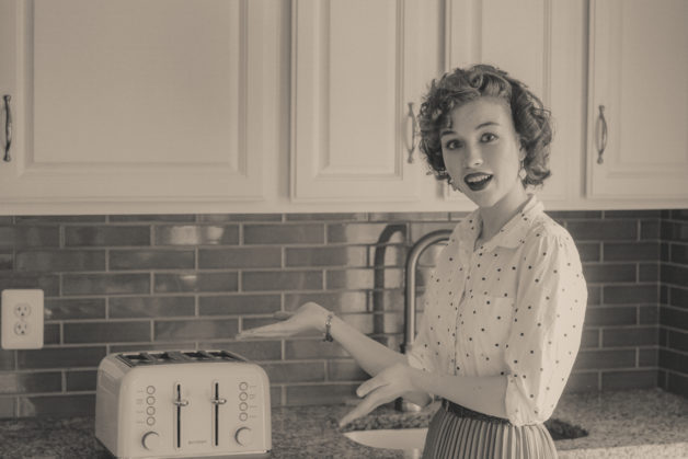 A black and white 1950s style photo of a curly-haired girl gesturing toward a toaster.