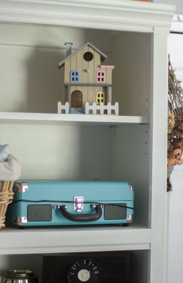 A white bookshelf with a birdhouse on the top shelf.