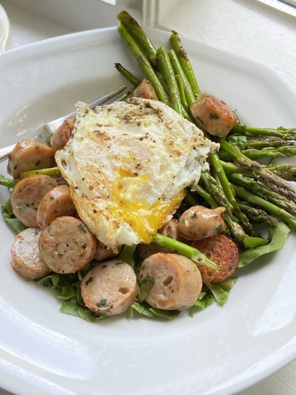 A white bowl with sausage, eggs, and asparagus inside of it.